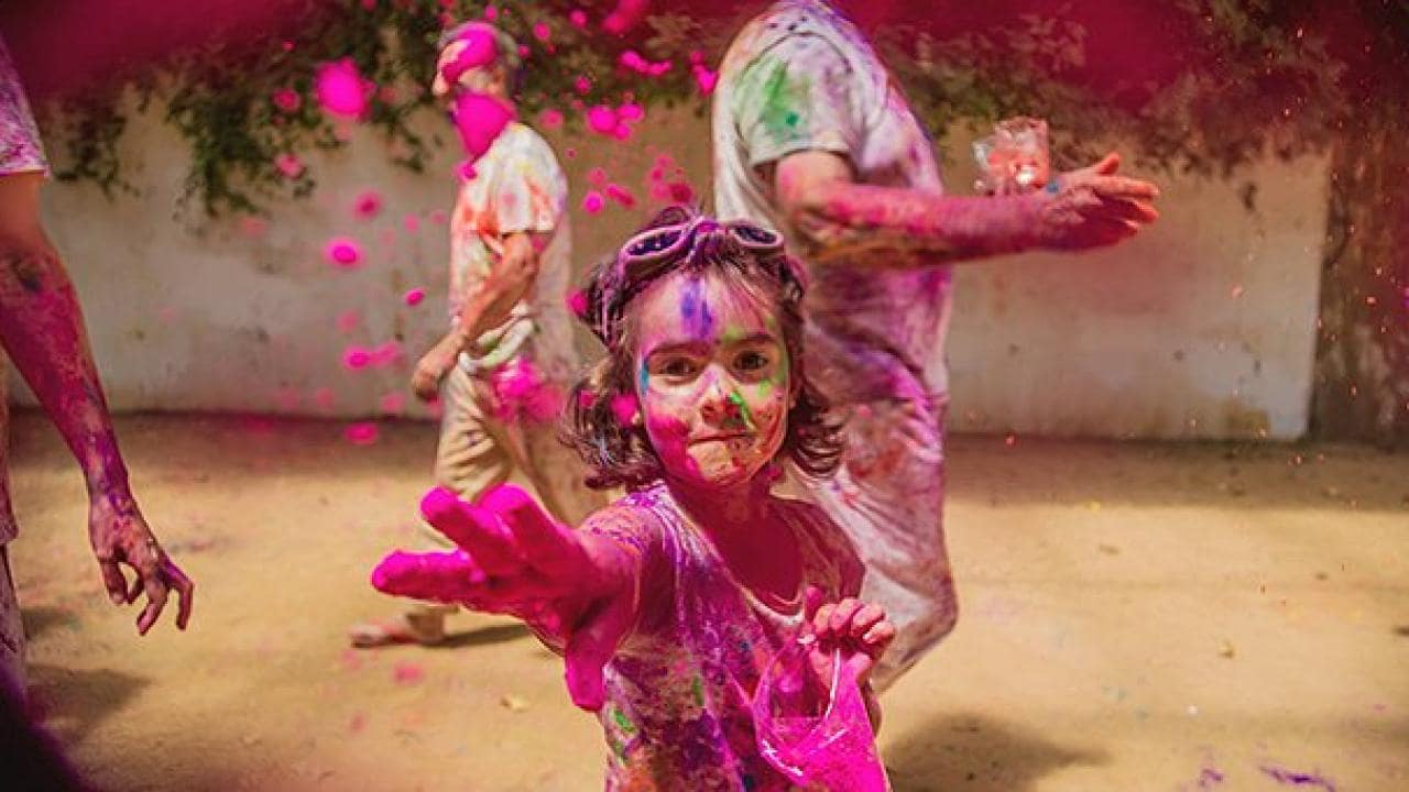 Holi kutlamaları sırasında renkli tozlara bulanmış bir çocuk kameraya doğru uzanıyor, arka plandaki diğer insanlar da canlı renklerle kaplanmış durumda.