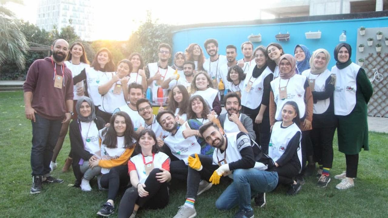 Eşleşen yelekler giyen bir grup insan çimenli bir alanda fotoğraf çektirmek için poz veriyor, gülümsüyor. Arka planda bir bina ve dekoratif duvar görünüyor.