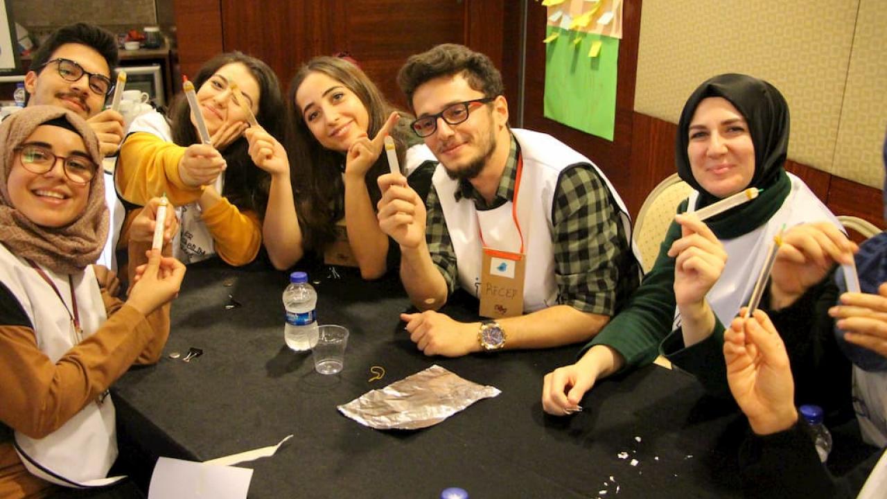 Bir masada oturan, üzerlerinde isim etiketleri ve ellerinde küçük eşyalar tutan bir grup insan kameraya gülümsüyor.