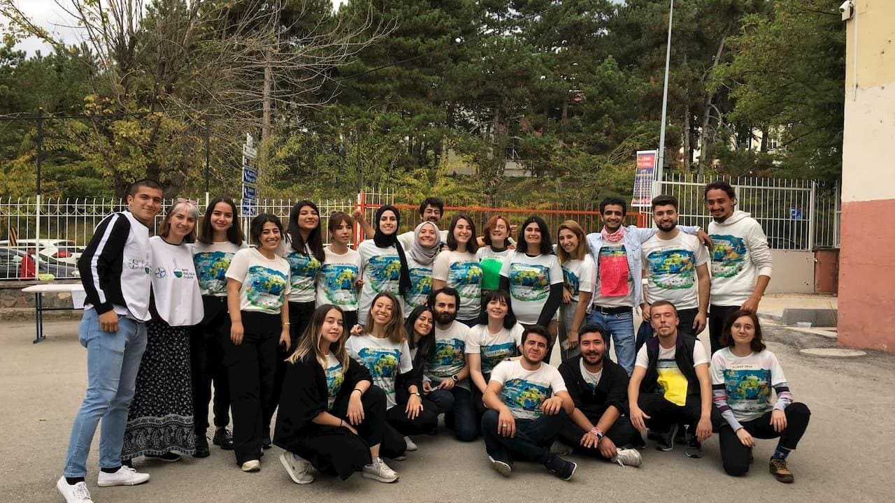 Aynı tişörtleri giyen bir grup insan, arka planda ağaçlar ve binalar varken açık havada birlikte poz veriyor.