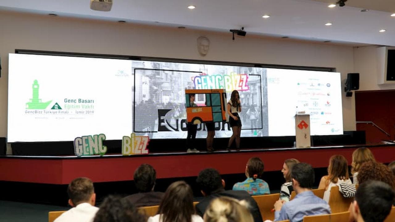 Bir konferansta sunum yapan insanlar, sahnede konuşmacılar ve arka planda bir ekran.
