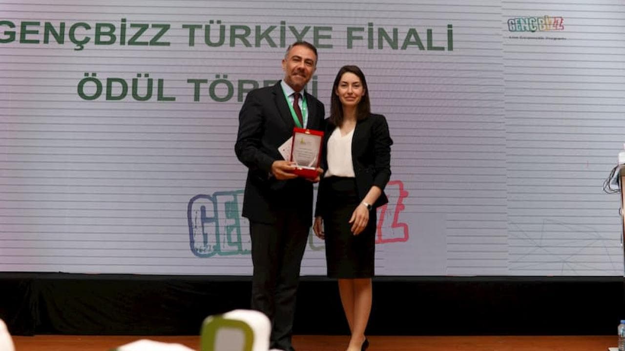 Sahnede iki kişi duruyor, birinin elinde ödül var. Arka planda logolu "GENÇBİZZ TÜRKİYE FİNALİ ÖDÜL TÖRENİ" yazısı yer alıyor.