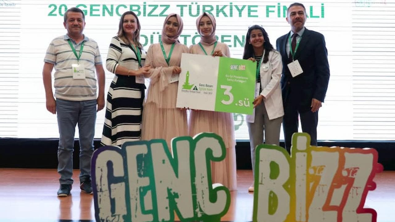 Altı kişilik bir grup sahnede duruyor ve ellerinde bir yarışma pankartı tutuyorlar, önlerinde ise "GENC BIZZ" yazılı bir pankart var.