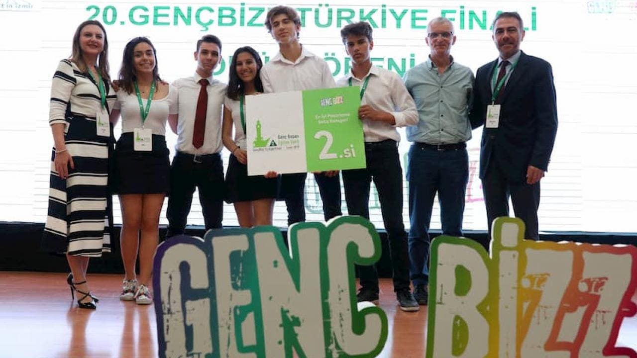 20. GençBizz Türkiye final etkinliğinde sahnede, üzerinde "2.si" yazılı bir pankart tutan bir grup genç yetişkin ve iki yaşlı adam.
