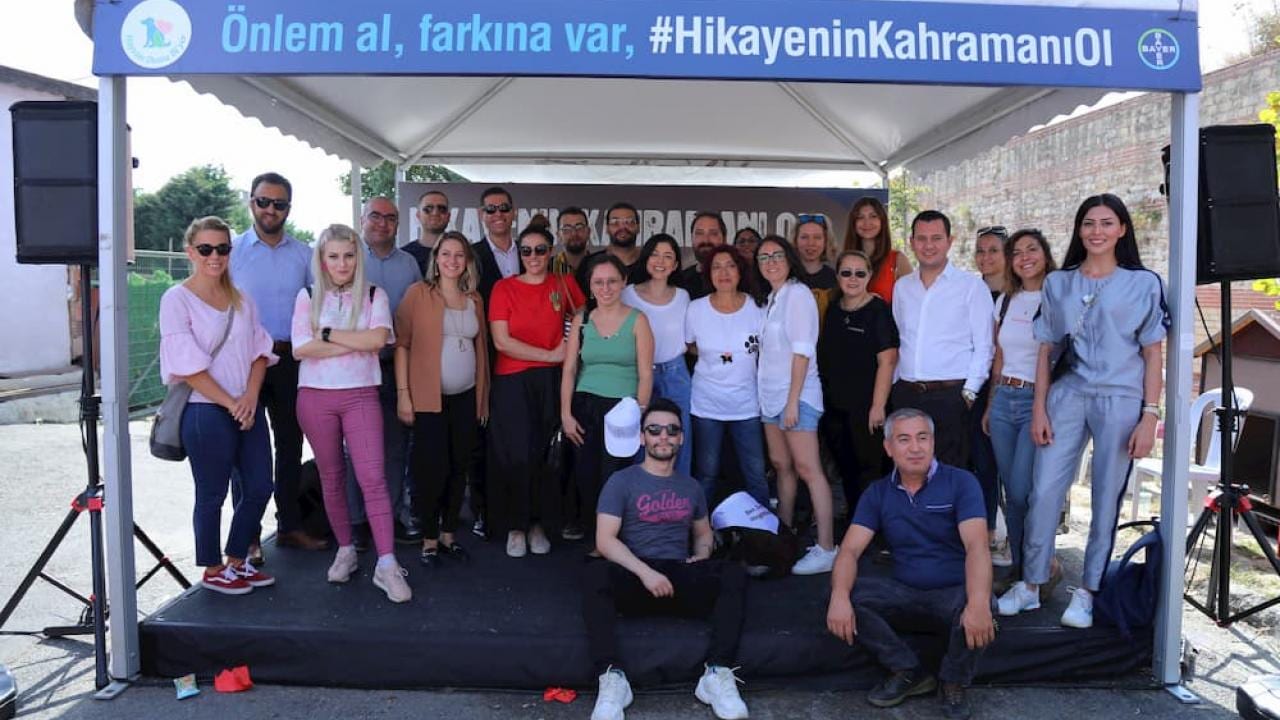 Üzerinde Türkçe yazılar bulunan pankartların bulunduğu bir çadırın altında birlikte poz veren bir grup insan.