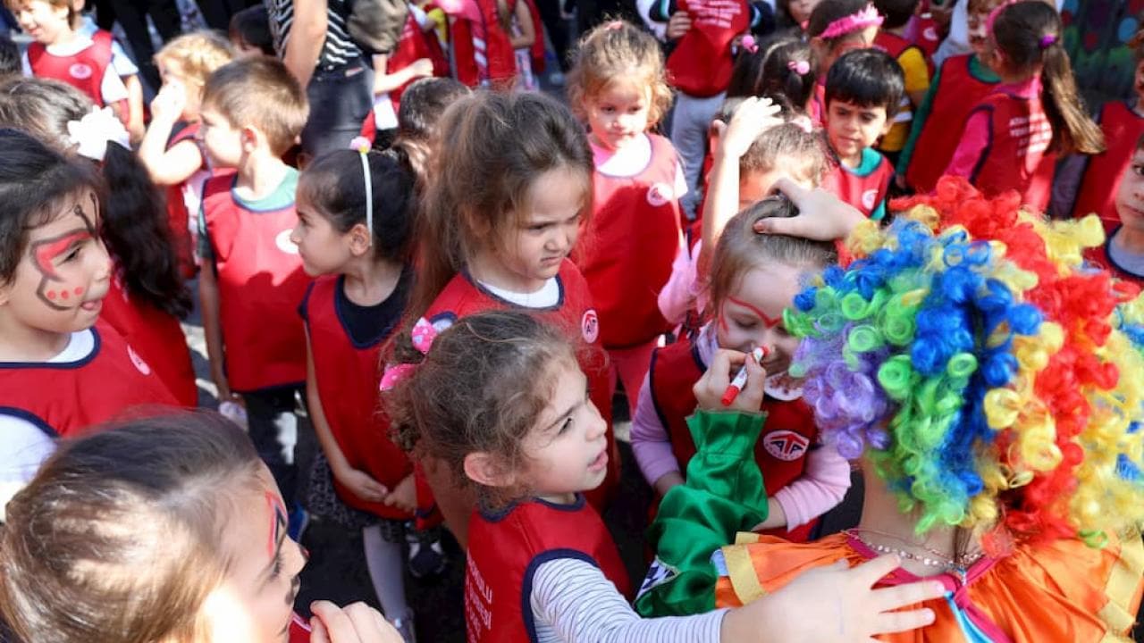 Kırmızı yelekli bir grup çocuk, açık hava etkinliğinde yüz boyayan bir palyaçonun etrafında toplanıyor.