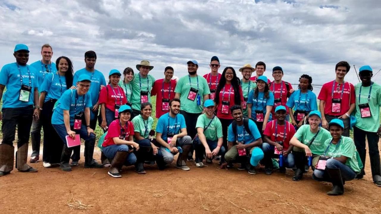 Group of people wearing colorful shirts and hats pose outdoors on a dirt path under a cloudy sky.
