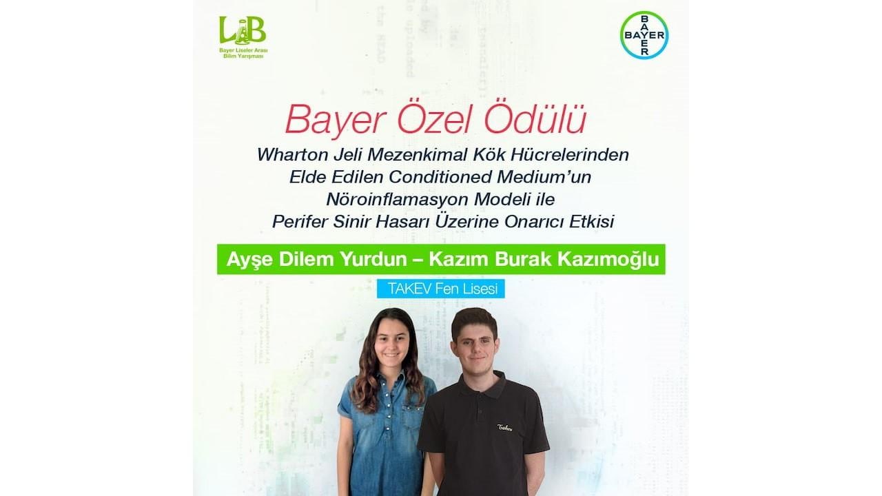 Bayer Özel Ödülü'nün sergilendiği panonun önünde iki kişi duruyor. Ödül açıklamasının altında isimleri Ayşe Dilem Yurdun ve Kazım Burak Kazımoğlu yer alıyor.