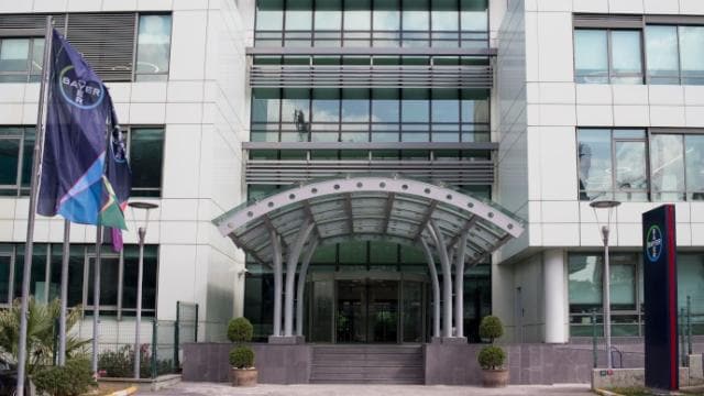 Modern cam kanopili, Bayer bayraklı ve yol kenarında saksı bitkileriyle donatılmış ofis binası girişi.