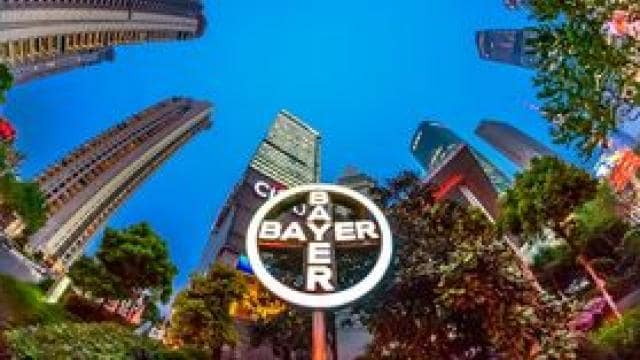 Ön planda ağaçlarla çevrili, açık mavi gökyüzüne karşı belirgin bir Bayer logosunun da yer aldığı kentsel gökdelenlerin balık gözü görünümü.