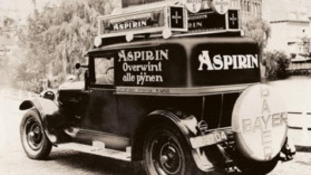 Üzerinde birden fazla dilde "Aspirin" reklamı bulunan ve arkasında "Bayer" yazan büyük bir stepne bulunan eski bir arabanın siyah beyaz görüntüsü.