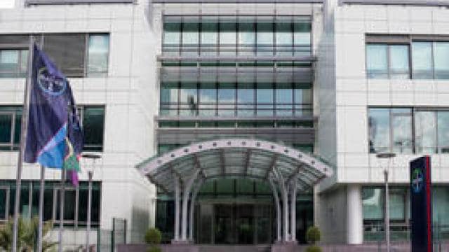 Cam pencereli modern bir ofis binasının ön görünümü. Girişin önünde iki bayrak görülüyor.